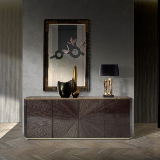 A modern, elegant room with the Contemporary Grey Buffet boasting a dark wooden sunburst pattern. Atop the buffet are two abstract vases, a metallic sculpture, and a black lampshade. Above the buffet hangs a large framed mirror reflecting contemporary art.