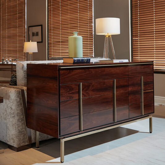 Contemporary Elegant Double Dresser A contemporary living room with a Contemporary Elegant Double Dresser featuring a dark, polished finish and sleek metal handles. On top are a green ceramic vase, a white book, and an elegant lamp with a clear base and white lampshade. The background showcases large windows with wooden blinds.