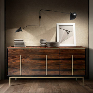 Contemporary Elegant Double Dresser A modern wooden sideboard with sleek brass handles stands against a neutral-toned wall. It holds a stack of books, several ceramic bowls, and a framed abstract artwork. A stylish wall-mounted black lamp with two adjustable arms illuminates the setup.