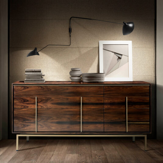 A modern wooden sideboard with sleek brass handles stands against a neutral-toned wall. It holds a stack of books, several ceramic bowls, and a framed abstract artwork. A stylish wall-mounted black lamp with two adjustable arms illuminates the setup.