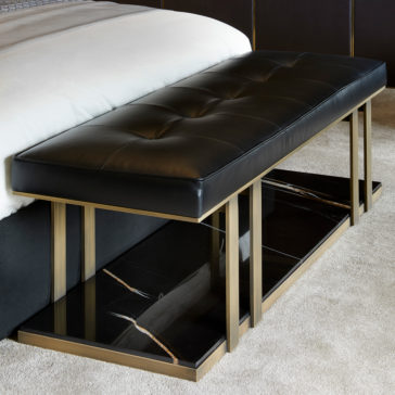 Contemporary Leather Bench The Contemporary Leather Bench, featuring a black tufted cushion and a brass frame with rectangular accents, is elegantly positioned at the foot of the bed. It also includes a black marble shelf underneath. The bench rests on a light beige carpeted floor in this modern bedroom.