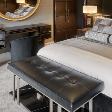 Contemporary Leather Bench A stylish bedroom features a modern gray bed adorned with decorative pillows, complemented by a Contemporary Leather Bench at the foot. The room also includes wooden side tables with lamps, a large round mirror above a sleek vanity paired with a black chair. The decor is minimalistic with neutral tones.