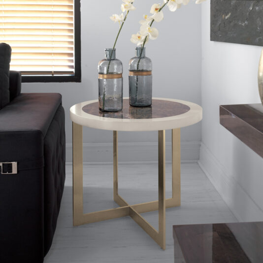 The Contemporary Marble Top Side Table with a white and maroon surface and gold legs is positioned next to a dark sofa. Two gray vases adorned with gold bands, each containing white orchid stems, sit on the table. In the background, there is a window with horizontal blinds.