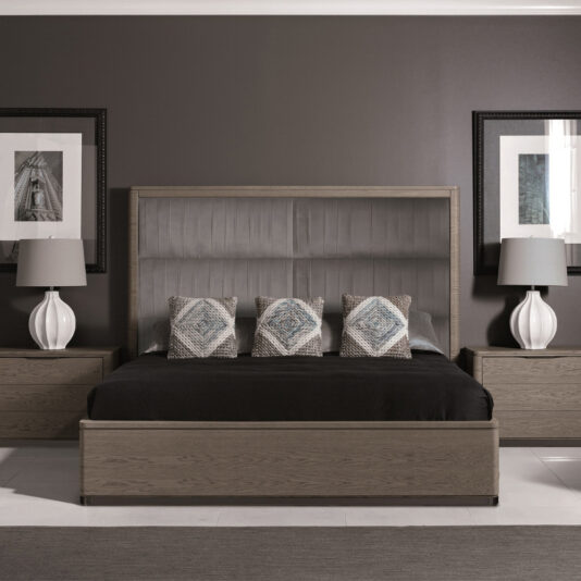 A modern bedroom with a dark gray accent wall features the Contemporary Oak Veneer Upholstered Bed, which boasts a tall, gray headboard and black bedding. The bed is adorned with four decorative pillows. Matching bedside tables with white lamps are positioned on either side, while framed artwork hangs above each table.