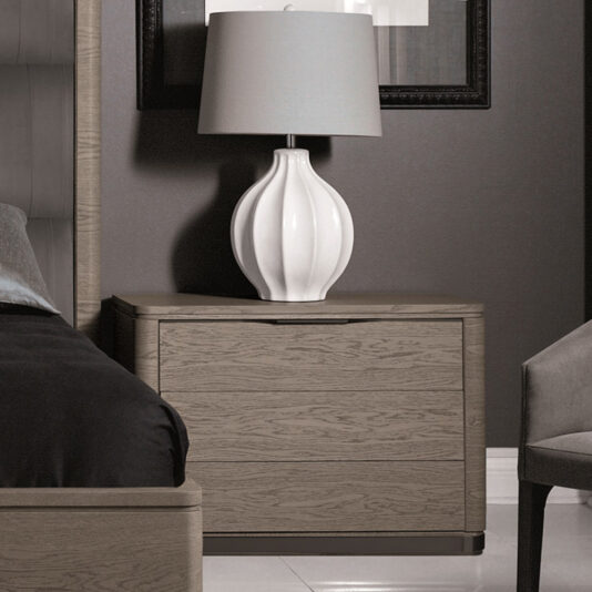 A modern bedroom nightstand, specifically called the Contemporary Oak Veneer Bedside Table, is paired with a large, white ceramic lamp featuring a textured, wavy design. The nightstand boasts a minimalist light wood finish with two drawers. Part of a bed with dark bedding and a grey chair can be seen on either side.