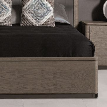 Contemporary Oak Veneer Upholstered Bed A stylish bedroom featuring a Contemporary Oak Veneer Upholstered Bed with a black bedspread. The bed is adorned with two accent pillows that have gray, white, and blue geometric patterns. A matching wooden nightstand with a round white lamp sits next to the upholstered bed.