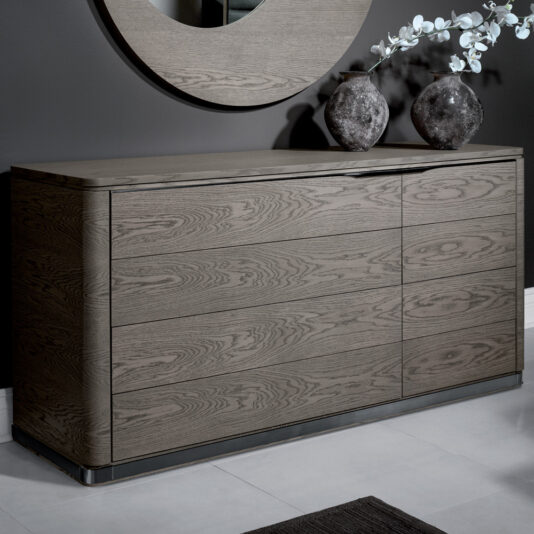 A sleek, modern Contemporary Oak Veneer Chest Of Drawers with a smooth, natural finish stands against a dark gray wall. Atop, two rustic vases with white flowers and a partially visible round mirror adorn the surface. The floor features light gray tile with a dark rug partially visible in the foreground—a true contemporary oak veneer chest of drawers ensemble.