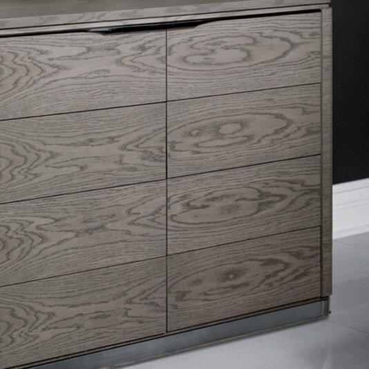 Contemporary Oak Veneer Chest Of Drawers The Contemporary Oak Veneer Chest of Drawers is a modern, light grey wooden dresser showcasing a smooth surface and natural wood grain patterns. This sleek design features five horizontal drawers, each with subtle groove handles. It stands gracefully on a slim, metallic base, perfectly complementing a minimalist room.