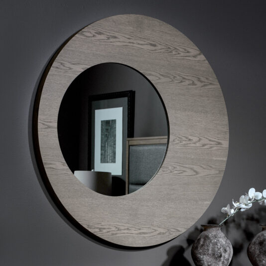 The Contemporary Oak Veneer Mirror, featuring a thick wooden frame, reflects part of a room that includes a framed picture on the wall and some furniture. On the table in the foreground are two decorative vases and a branch with white flowers. The dark gray wall behind the mirror contrasts beautifully with its design.