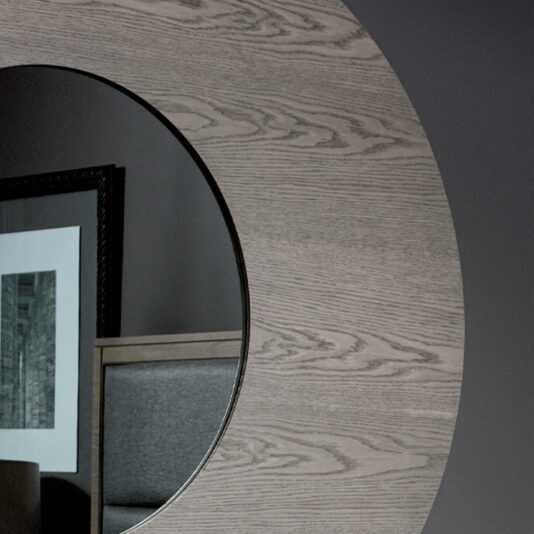 A close-up view of the Contemporary Oak Veneer Mirror reflecting part of a modern interior. The reflection shows a framed black-and-white artwork on a gray wall and a portion of a piece of furniture with a gray upholstered backrest.