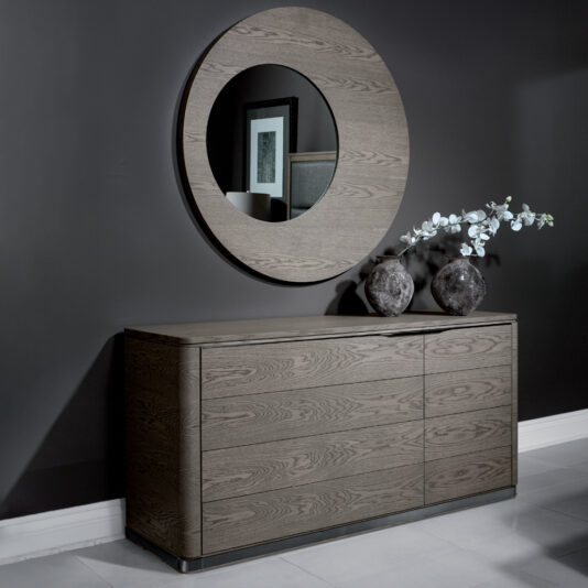 A modern interior featuring a wooden sideboard with a sleek design against a dark gray wall. Above the sideboard hangs a large round mirror. Two decorative vases with white flowers sit atop the sideboard, and a framed picture is partially reflected in the mirror.