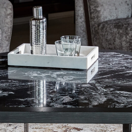 A close-up of the Contemporary Octagonal Coffee Table with a white tray holding a metal cocktail shaker and two clear glasses. Two plush, grey armchairs are partially visible in the background. The table's surface has a glossy, multicolored marble pattern.