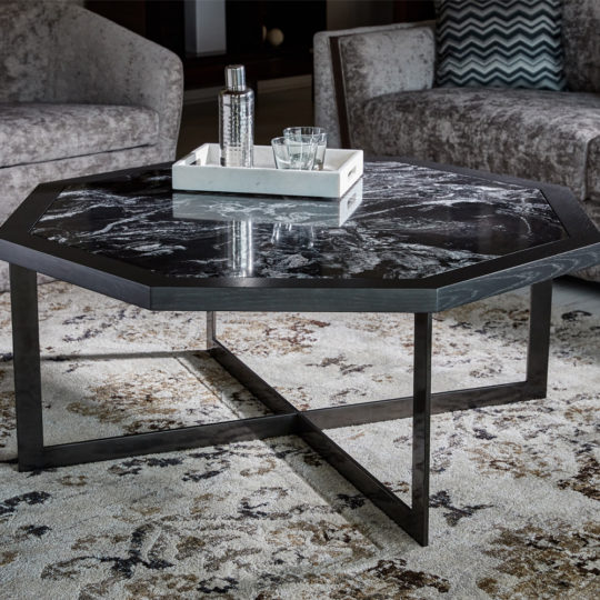 A modern living room features the Contemporary Octagonal Coffee Table, crafted from sleek black marble and supported by a stylish metal frame. On the table, a white tray holds a silver flask and two glasses. The setting is completed with contemporary gray chairs and a patterned area rug.