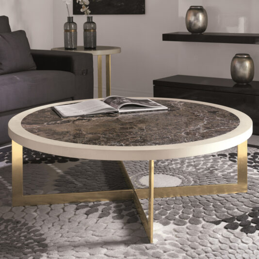 A stylish living room showcases the Contemporary Round Marble Top Coffee Table with elegant gold metal legs. An open book rests on the table. In the background, a side table adorned with vases sits next to a grey couch. The floor features a textured patterned rug in shades of grey.