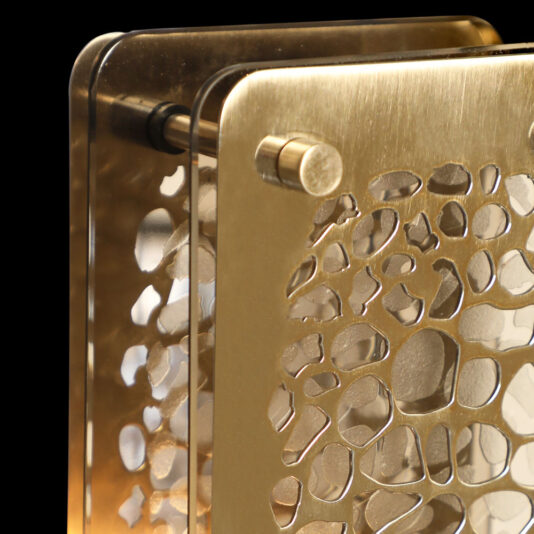 Contemporary Marble Table Lamp A close-up of the contemporary marble table lamp reveals its sleek, modern metallic design and a distinctive, intricate cutout pattern. The lamp features a golden hue and an elegant layered structure, with two flat, rectangular pieces connected by small cylindrical rods. Set against a black background, the lamp's elegance is further accentuated.