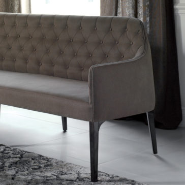 Contemporary Three Seater Sofa A gray, upholstered Contemporary Three Seater Sofa with a tall, tufted backrest and sleek, dark wooden legs sits on a light-colored floor next to a curtain. This modern piece boasts a subtle and elegant appearance.