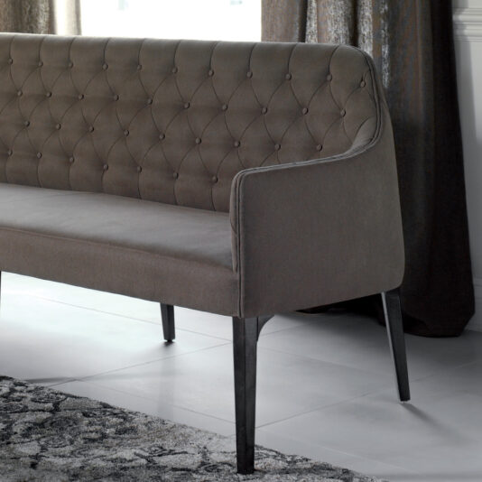 Contemporary Three Seater Sofa A gray, upholstered Contemporary Three Seater Sofa with a tall, tufted backrest and sleek, dark wooden legs sits on a light-colored floor next to a curtain. This modern piece boasts a subtle and elegant appearance.