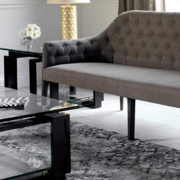 Contemporary Three Seater Sofa A modern living room scene featuring the Contemporary Three Seater Sofa with black wooden legs, a glass coffee table, and an end table. On the end table are a gold textured lamp and a vase with white flowers. The room has a gray and white patterned rug on a tiled floor.