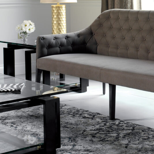 Contemporary Three Seater Sofa A modern living room scene featuring the Contemporary Three Seater Sofa with black wooden legs, a glass coffee table, and an end table. On the end table are a gold textured lamp and a vase with white flowers. The room has a gray and white patterned rug on a tiled floor.