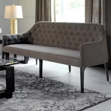 Contemporary Three Seater Sofa A modern living room features the Contemporary Three Seater Sofa in dark gray, with slender black legs. The sofa is placed on a gray patterned rug, adjacent to a sleek glass coffee table with a black frame. A gold lamp with a white shade stands on a side table near the curtained window.
