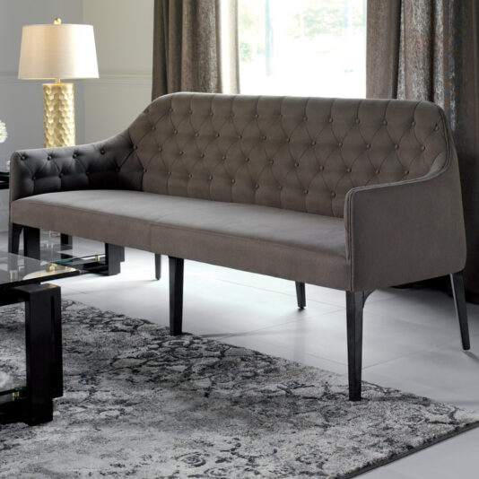 A modern living room features the Contemporary Three Seater Sofa in dark gray, with slender black legs. The sofa is placed on a gray patterned rug, adjacent to a sleek glass coffee table with a black frame. A gold lamp with a white shade stands on a side table near the curtained window.