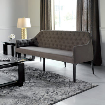 Contemporary Three Seater Sofa A modern living room featuring a Contemporary Three Seater Sofa with gray tufted upholstery and black legs. The room includes a glass coffee table with a black frame, a side table with a gold-textured lamp, and long, dark curtains hanging from the window. The floor has a sleek, patterned rug.