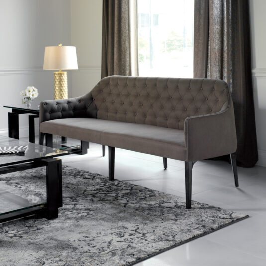 Contemporary Three Seater Sofa A modern living room featuring a Contemporary Three Seater Sofa with gray tufted upholstery and black legs. The room includes a glass coffee table with a black frame, a side table with a gold-textured lamp, and long, dark curtains hanging from the window. The floor has a sleek, patterned rug.