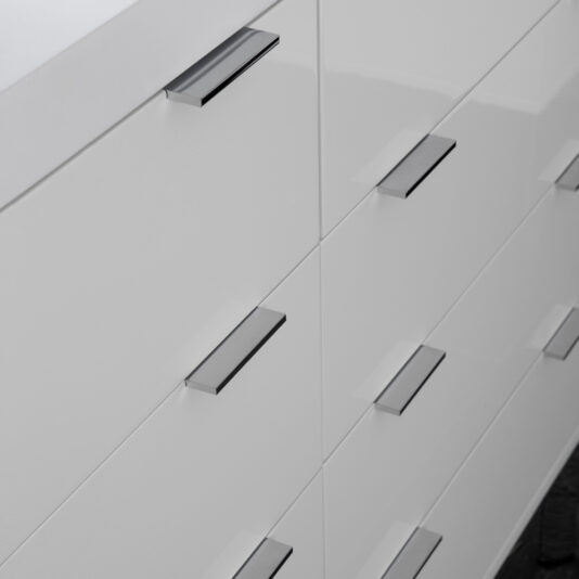 Contemporary Miami Style White Double Dresser Close-up of the Contemporary Miami Style White Double Dresser, showcasing sleek, minimalistic metal handles on its drawers. The clean lines and glossy finish enhance the modern design, with an angled perspective emphasizing the dresser's symmetry and simplicity.