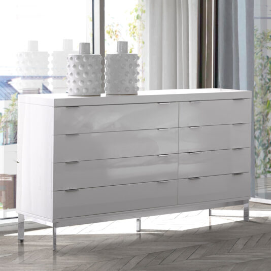 The Contemporary Miami Style White Double Dresser with six drawers stands in front of a large window. The dresser features sleek, minimalist handles and metallic legs. Two white, textured vases are placed on top. Sunlight streams through the window, illuminating the room.