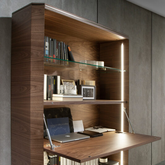 Contemporary Wooden Writing Desk The showcased Contemporary Wooden Writing Desk features an extended writing surface and integrated shelving lined with books, a laptop, and decorative items. The desk includes built-in lighting, creating a warm and functional workspace within a minimalist setting.