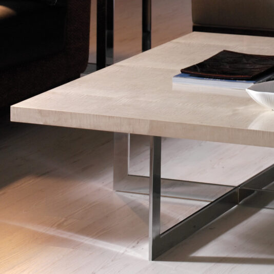 A modern living room with a Contemporary Wooden Coffee Table featuring a sleek metal frame. On the table, there are a few books stacked and a white bowl. The floor has a light wood finish and part of a brown sofa is visible in the background.