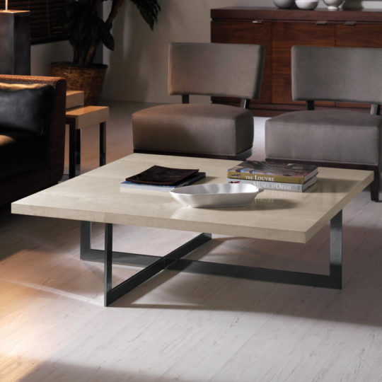 A modern living room showcases the Contemporary Wooden Coffee Table With Black Base, adorned with a white decorative bowl, a book, and a magazine. In the background, two grey armchairs and a wooden sideboard complement the scene. The light-colored flooring completes the stylish look.