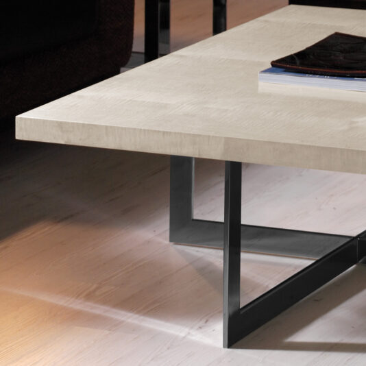 A close-up of the Contemporary Wooden Coffee Table With Black Base showcases its light-colored tabletop and sleek black base. On top of the table, a few stacked magazines or books add to its minimalistic charm. The surrounding floor and part of a dark-colored sofa are partially visible in the background.