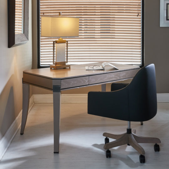 A modern home office with a Contemporary Wooden Writing Desk under a window with closed blinds. A stylish black swivel chair with wooden legs is placed at the desk, which features an open book and a contemporary table lamp, casting a warm light.