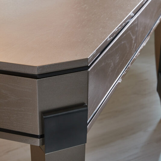 A close-up of the corner of the Contemporary Wooden Writing Desk reveals its sleek, modern design with a slightly textured, metallic surface. The desk showcases clean lines and a minimalist aesthetic, supported by dark rectangular legs that enhance its overall contemporary appeal.