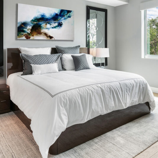 A deluxe contemporary bedroom features the Deluxe Contemporary Veneered Bed adorned with white linens and gray accent pillows. Above the bed, a colorful abstract painting adds a splash of vibrant color. The room has a floor-to-ceiling window on the right, allowing natural light to flood the space. A lamp sits on a bedside table next to the bed.