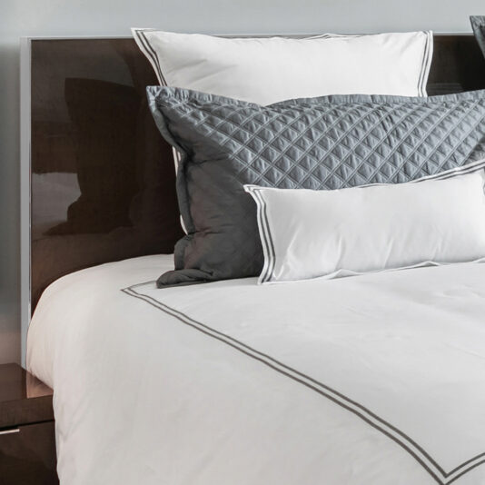 A neatly made bed showcasing a Deluxe Contemporary Veneered Bed, adorned with a white comforter and pillows. It includes a decorative quilted gray pillow and a smaller white pillow with gray borders, all set against an elegant wooden headboard.