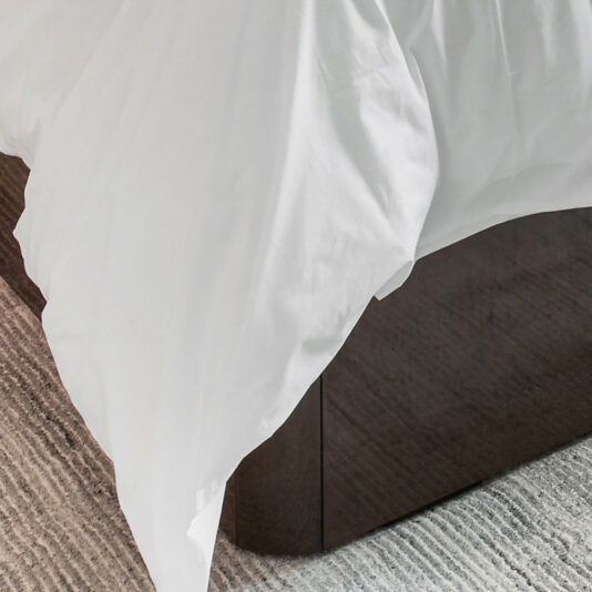 A neatly made bed with white sheets and a visible dark wooden frame is shown. This Deluxe Contemporary Veneered Bed is placed on a textured, light-colored carpet. The white sheets hang slightly over the edges of the bed.