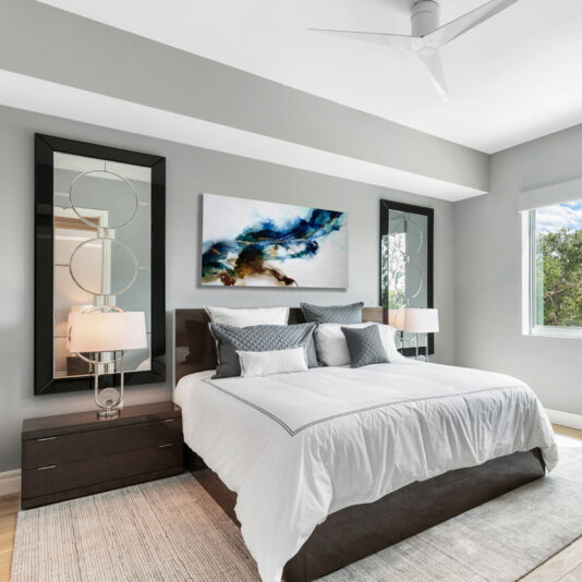 A deluxe contemporary bedroom featuring a neatly made Deluxe Contemporary Veneered Bed with white bedding and several gray and white pillows. Two dark wooden nightstands with matching lamps flank the bed. Above it hangs a colorful abstract painting, and two large mirrors are on either side. A window lets in natural light.