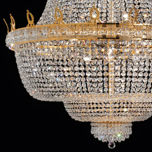 A close-up shot showcases an intricate Deluxe Crystal Drop Empire Chandelier against a black background. The chandelier boasts elaborate gold detailing and numerous sparkling crystal beads, arranged in a cascading, tiered design. Bright light reflects off the crystals, creating a dazzling effect.