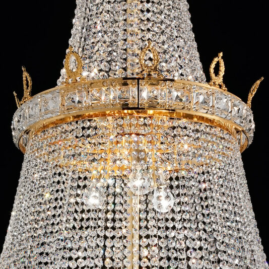 A close-up view of the Deluxe Crystal Drop Empire Chandelier reveals its elegant gold frame adorned with intricate designs and cascading patterns of sparkling crystal beads. The crystals reflect light, creating a dazzling and ornate display.