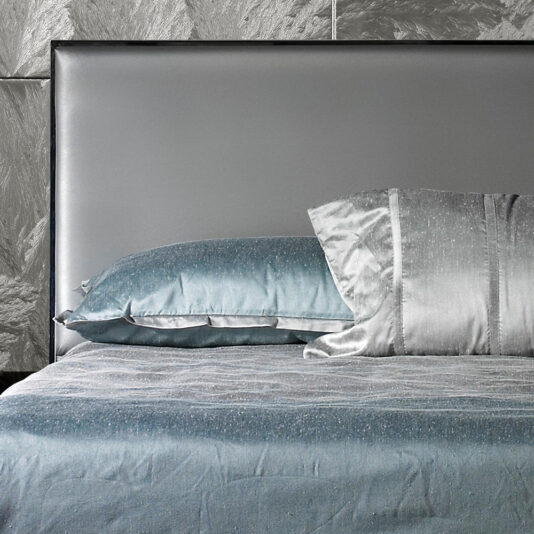The Deluxe Grey Leather Veneered Bed, featuring a sleek, upholstered headboard, sets the stage. It's adorned with a light blue sparkly blanket and coordinated pillows—one light blue and one silver. The background wall showcases a textured gray finish.