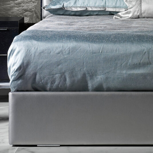 Close-up of a neatly made bed with a metallic gray bedspread and matching pillows. The padded and upholstered footboard and side of the Deluxe Grey Leather Veneered Bed are covered in smooth gray fabric. A portion of a dark wooden nightstand is visible on the left side of the image.
