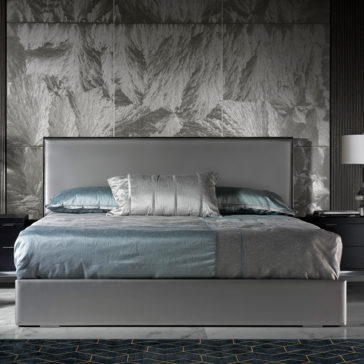 Deluxe Grey Leather Veneered Bed A modern bedroom features a Deluxe Grey Leather Veneered Bed with a tall headboard, dressed in gray and teal bedding with matching pillows. On either side of the bed are sleek black nightstands topped with white table lamps. The backdrop is a textured, silver-toned wall.