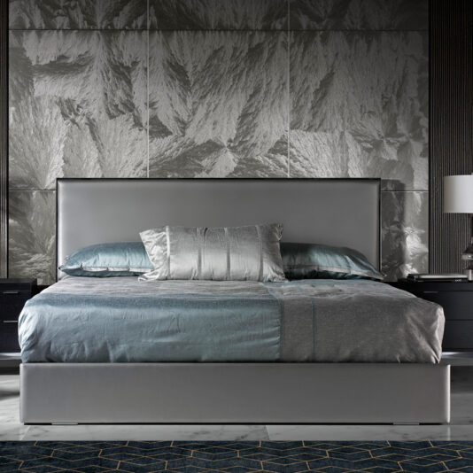 A modern bedroom features a Deluxe Grey Leather Veneered Bed with a tall headboard, dressed in gray and teal bedding with matching pillows. On either side of the bed are sleek black nightstands topped with white table lamps. The backdrop is a textured, silver-toned wall.