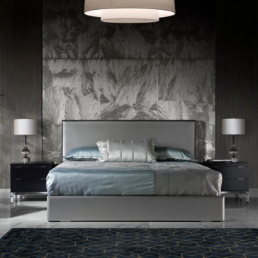 Deluxe Grey Leather Veneered Bed Modern bedroom featuring the Deluxe Grey Leather Veneered Bed, adorned with a mix of light grey and teal bedding. Two dark nightstands with lamps flank the bed, set against a textured metallic wall. A geometric-patterned area rug adorns the floor, completing this sophisticated space.