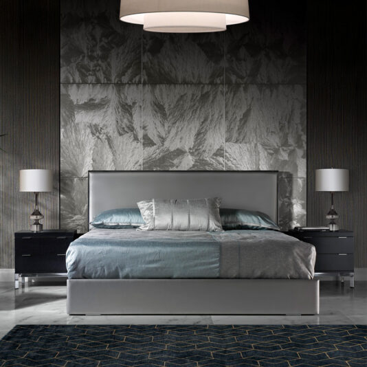 Modern bedroom featuring the Deluxe Grey Leather Veneered Bed, adorned with a mix of light grey and teal bedding. Two dark nightstands with lamps flank the bed, set against a textured metallic wall. A geometric-patterned area rug adorns the floor, completing this sophisticated space.