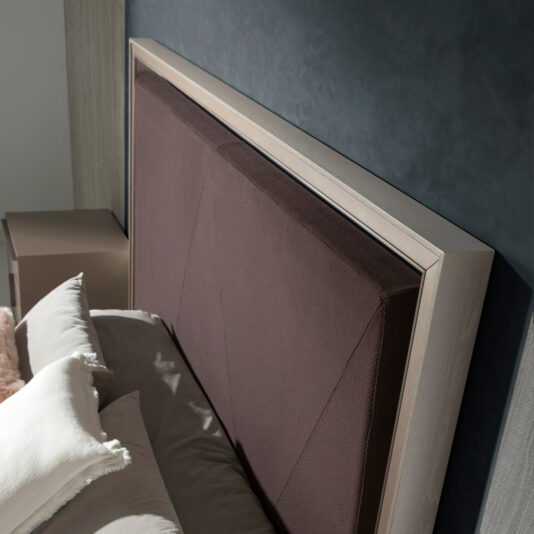 Close-up of a stylish bed with a fabric-upholstered headboard, featuring a geometric design. The headboard is framed with a light wood border. The bed is dressed with beige and white pillows and a light gray duvet, positioned against a dark gray wall.