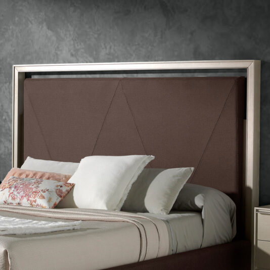 The Deluxe Modern Upholstered King Size Bed, featuring a rectangular, padded headboard in brown fabric, is beautifully adorned with an assortment of pillows - a white pillow, a white pillow with floral patterns, and beige pillows. The bed is elegantly set against a gray textured wall.