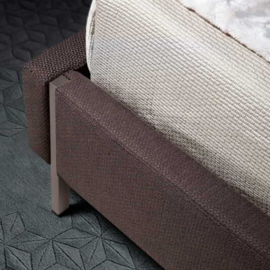 A close-up of a bed's corner, featuring a textured brown fabric frame, metallic legs, and a beige quilted blanket covering part of the mattress. The bed is placed on a dark, patterned carpet.